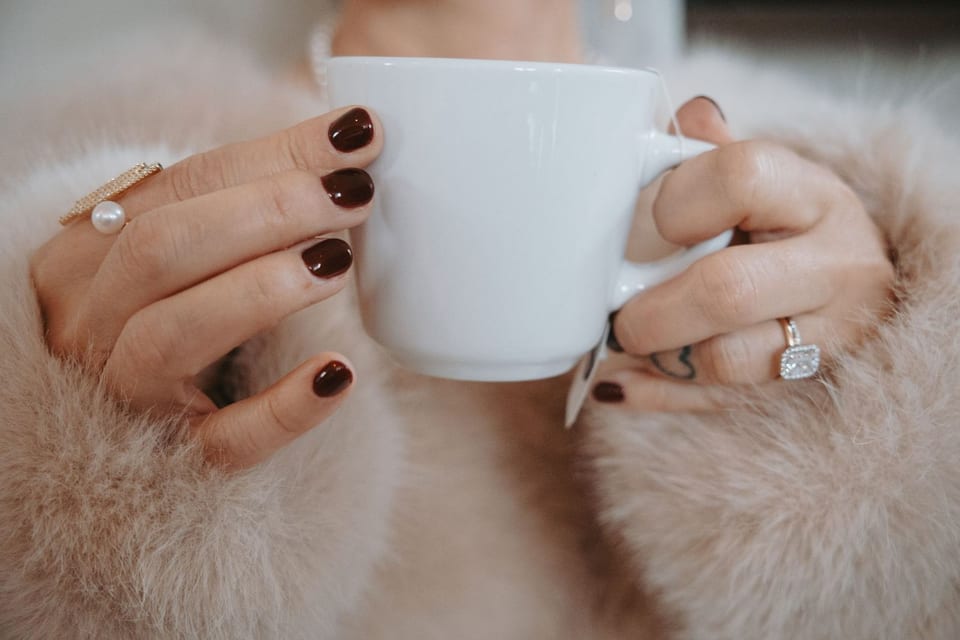 Close-up of the bride's hands wrapped in a fur stole, holding a coffee mug — engagement ring and pearl ring in frame.