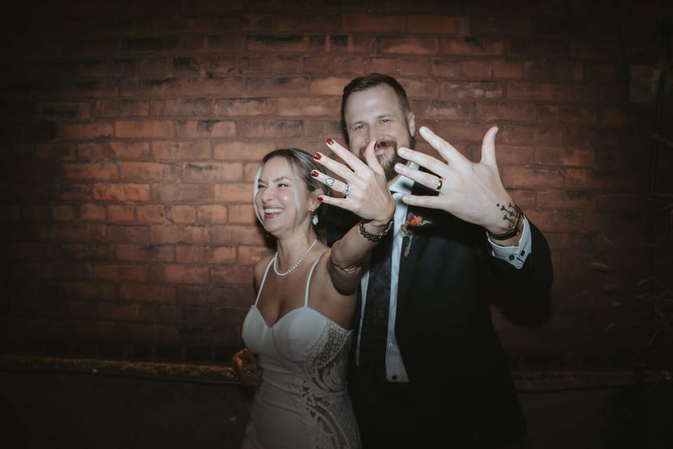 Newlyweds laughing as they show off their wedding rings against a warm brick wall.