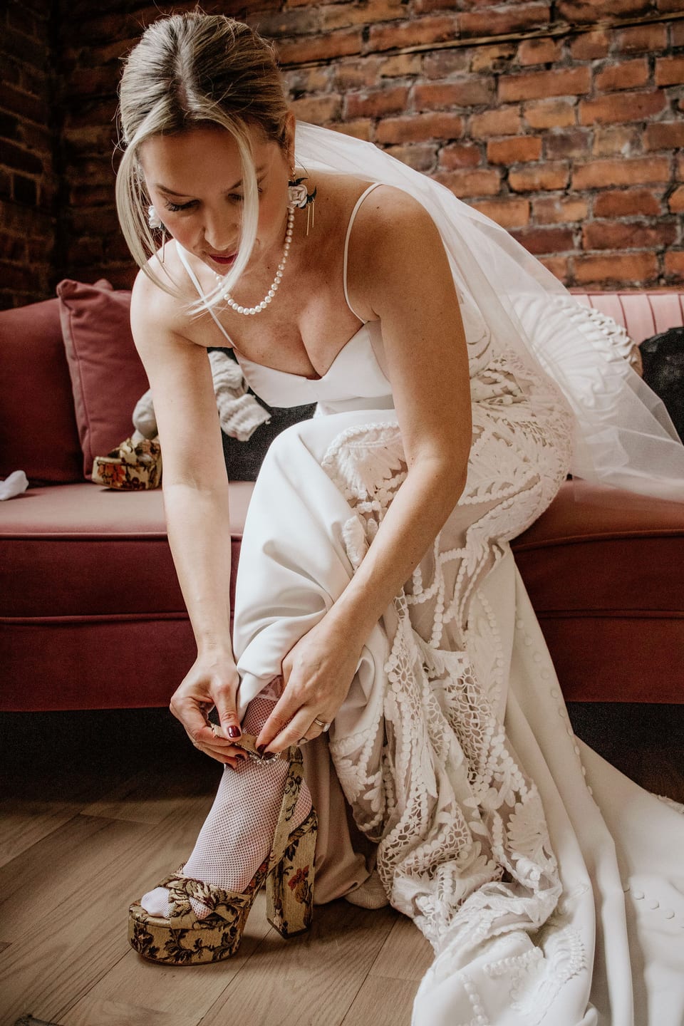 Bride fastening her shoe as she gets ready, seated on a velvet couch.