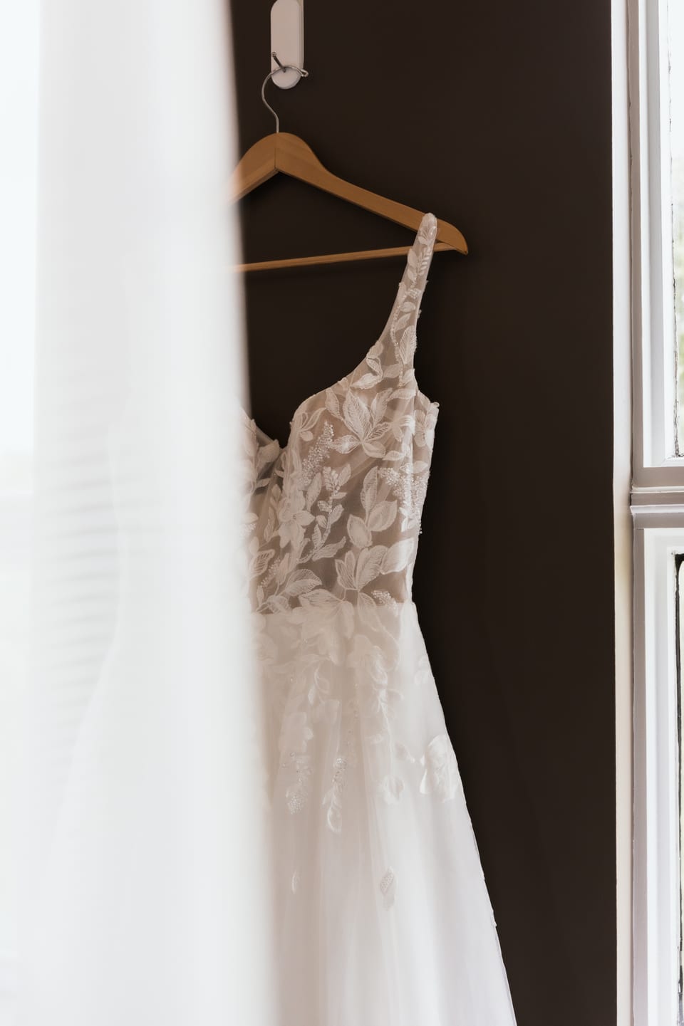 A one-shoulder floral lace wedding gown hangs from a wooden hanger against a dark wall, sheer curtain in the foreground.
