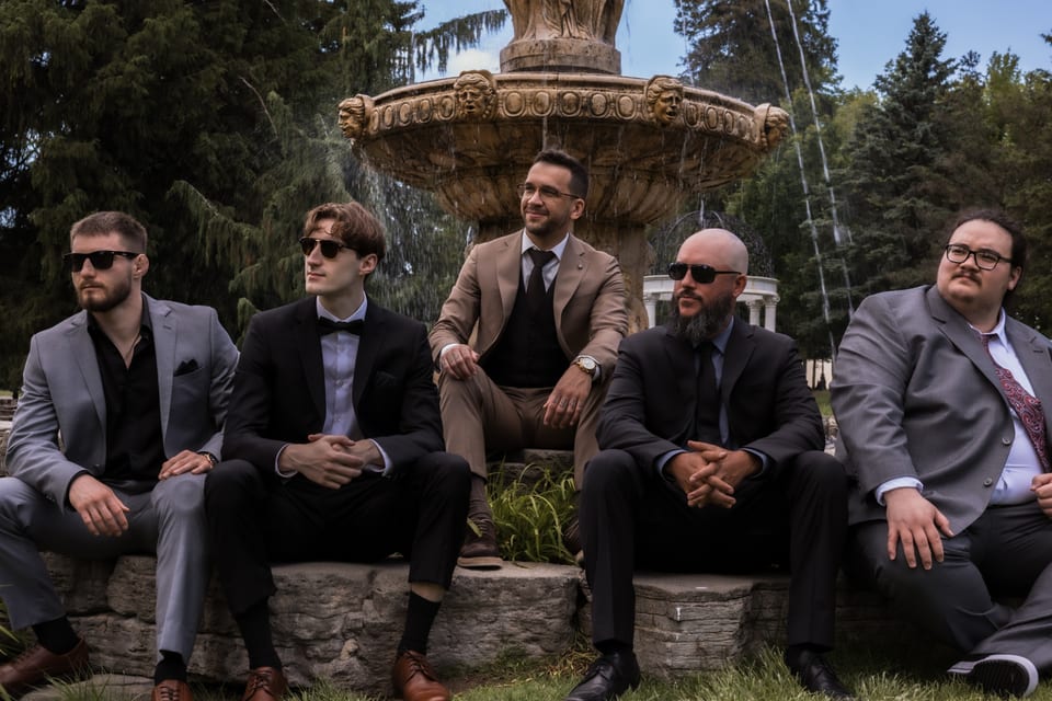 Five groomsmen in grey, brown, and black suits lounge on the stone steps of an ornate fountain, most wearing sunglasses.