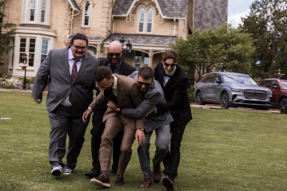A jubilant group of groomsmen carry the groom across the lawn in front of a grand brick house, laughing mid-stride.