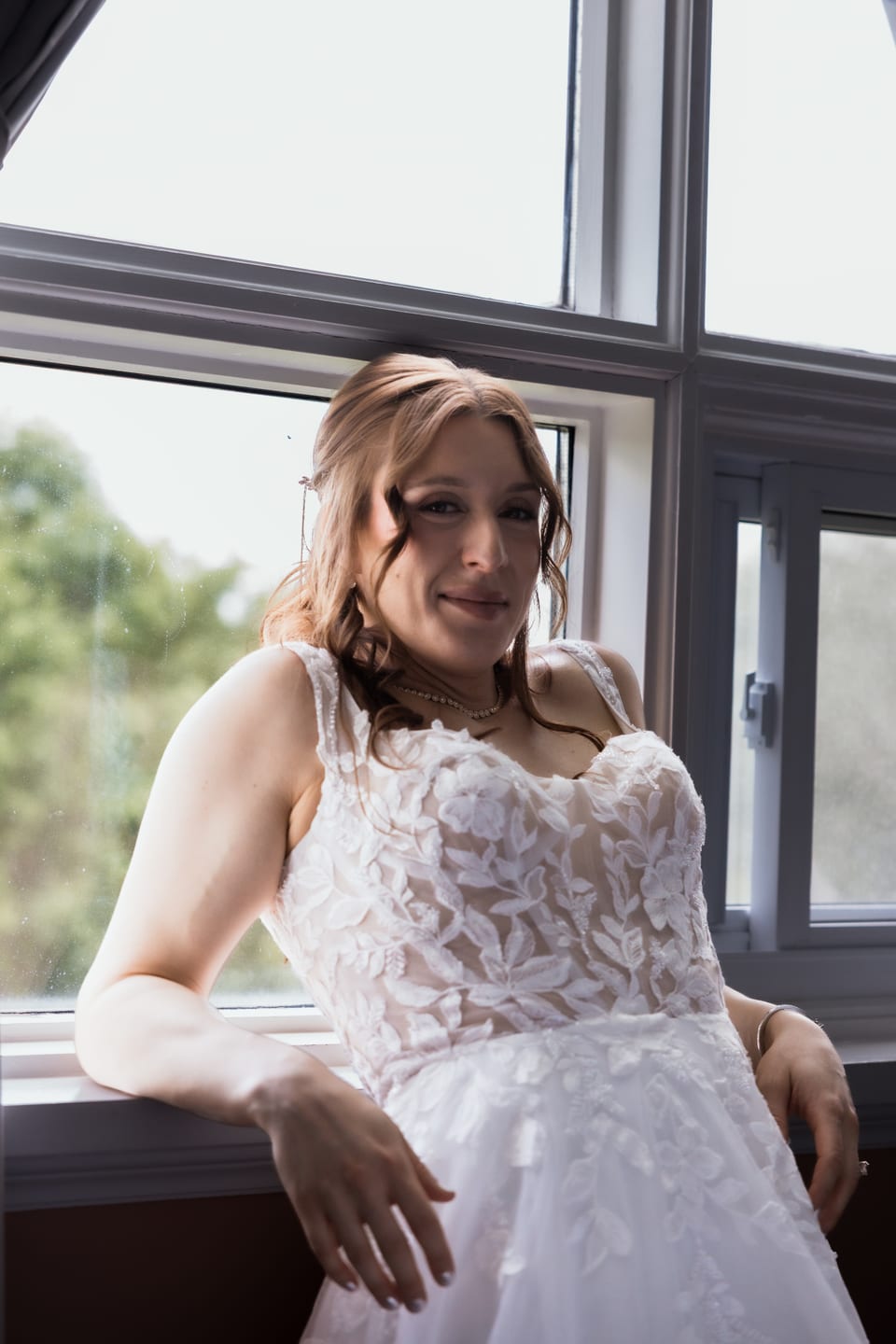 The bride leans against a window sill in her lace gown, smiling softly with a pearl choker and loose waves.
