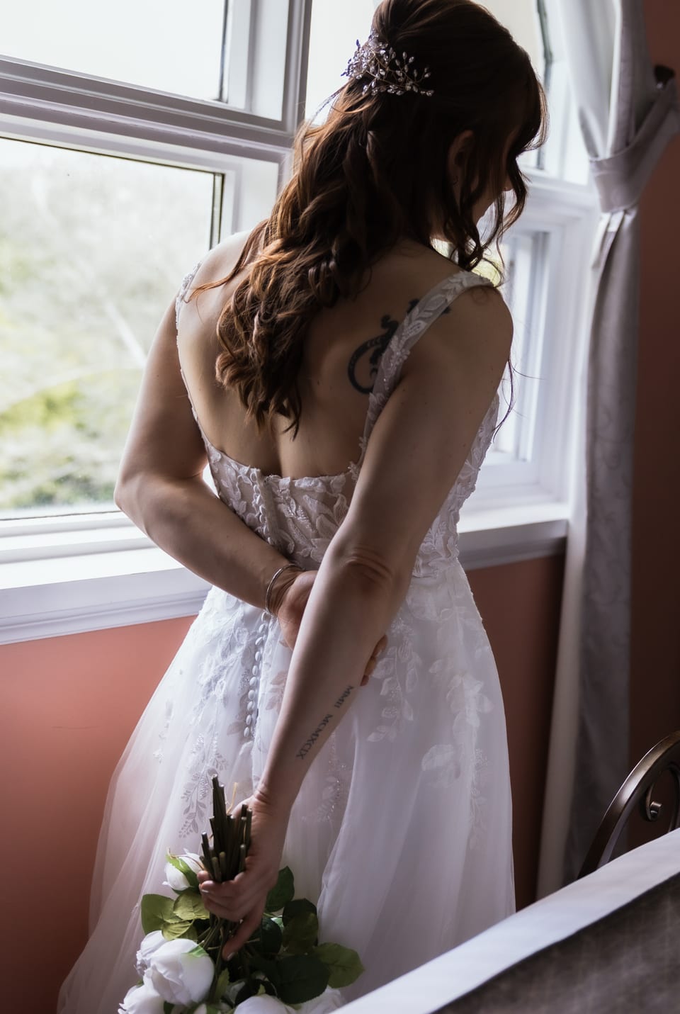 The bride's back tattoo and forearm ink as she's buttoned into her lace gown by a window, bouquet in hand.