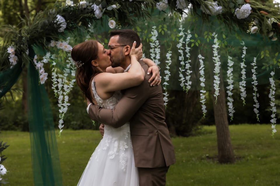 The bride and groom share a kiss beneath a floral ceremony arch woven with greenery and hanging white blossoms.