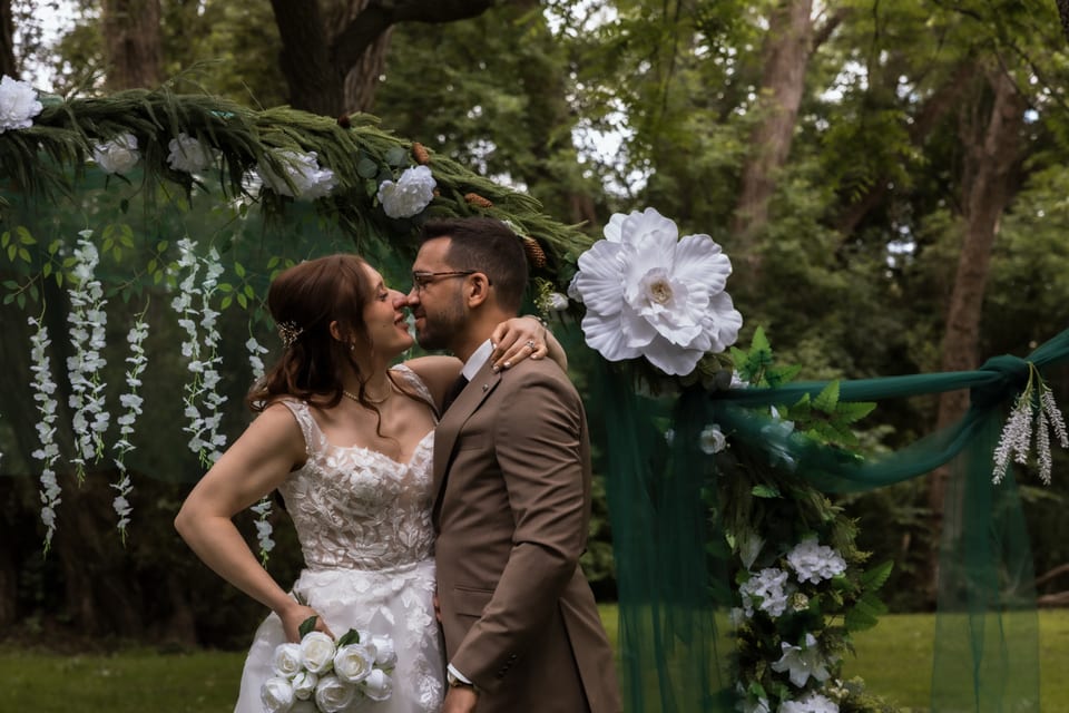 Bride and groom touch noses and smile under the floral ceremony arch, her bouquet of white roses in hand.