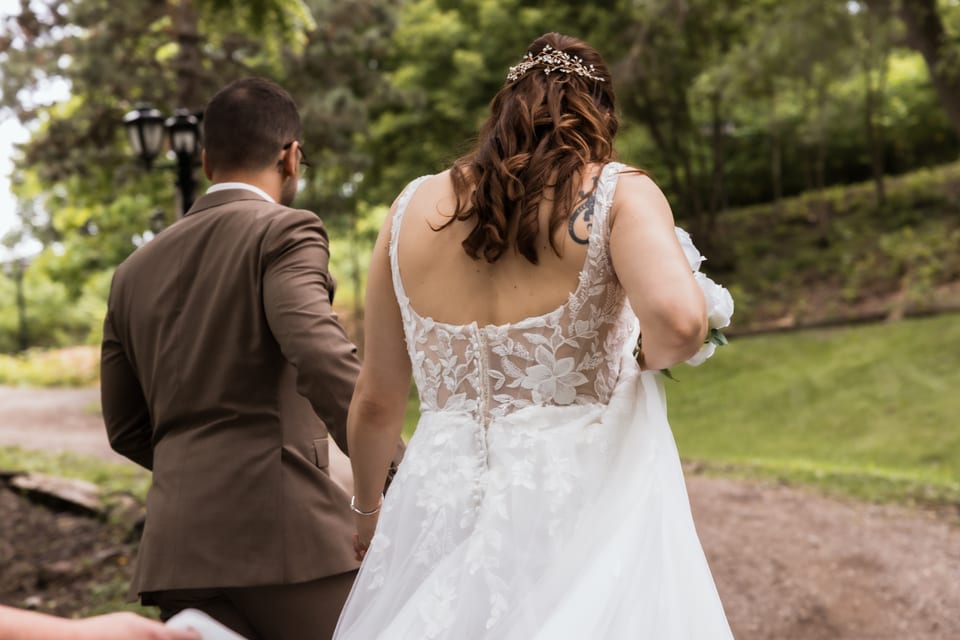 The couple walk hand-in-hand away from the ceremony down a garden path, her lace back and bouquet trailing.