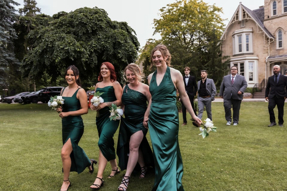 Four bridesmaids in emerald satin gowns laugh and walk across the lawn in front of a historic mansion.