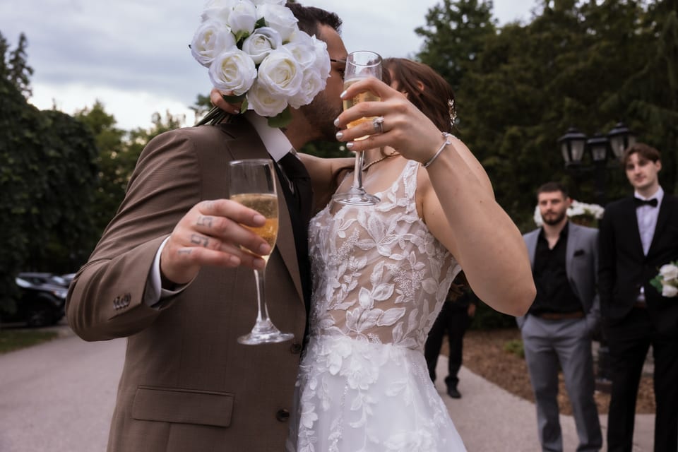 Bride and groom toast with champagne flutes, her bouquet of white roses held up between them.