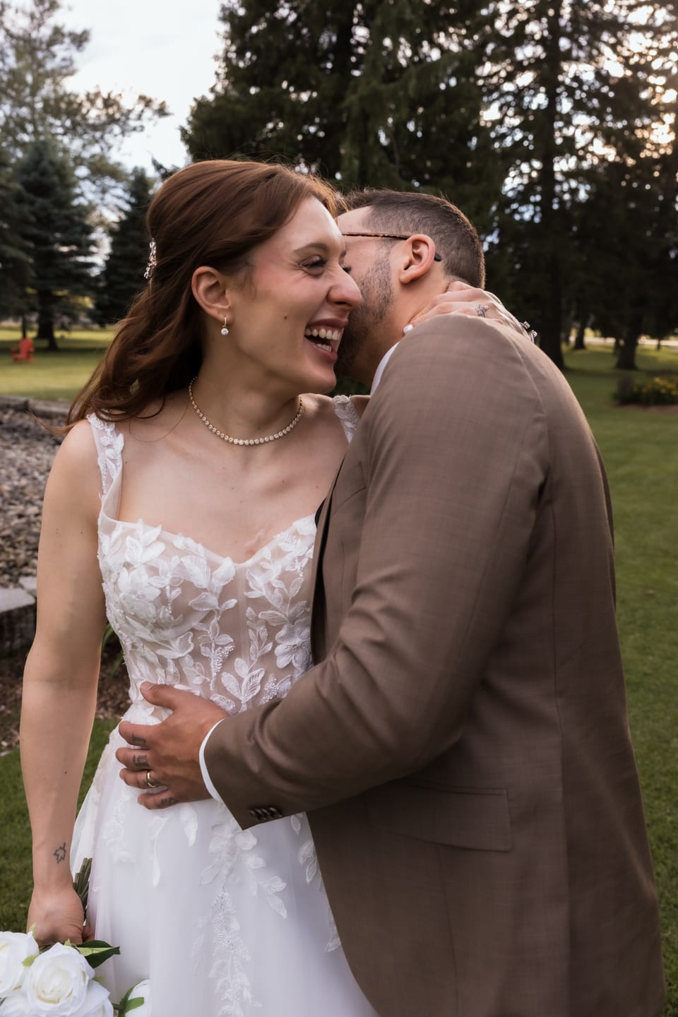 The bride laughs as the groom nuzzles her cheek, her diamond necklace and earrings catching the light.