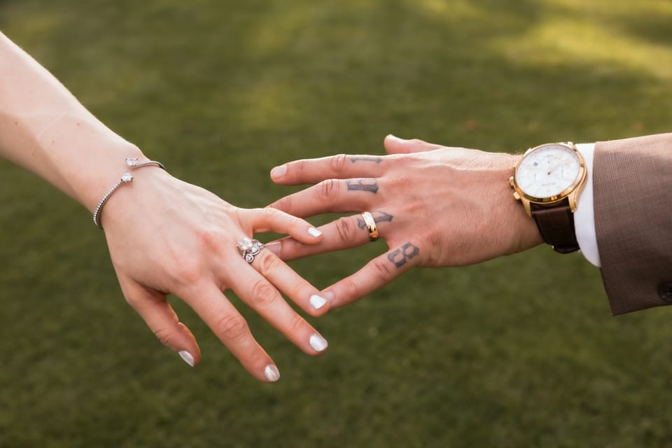 Close-up of the couple's hands reaching for each other, showing the engagement ring, wedding band, and tattoos.