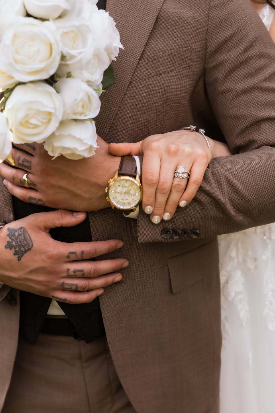 Detail of the couple's embracing hands and rings resting against the groom's brown suit, bouquet beside them.