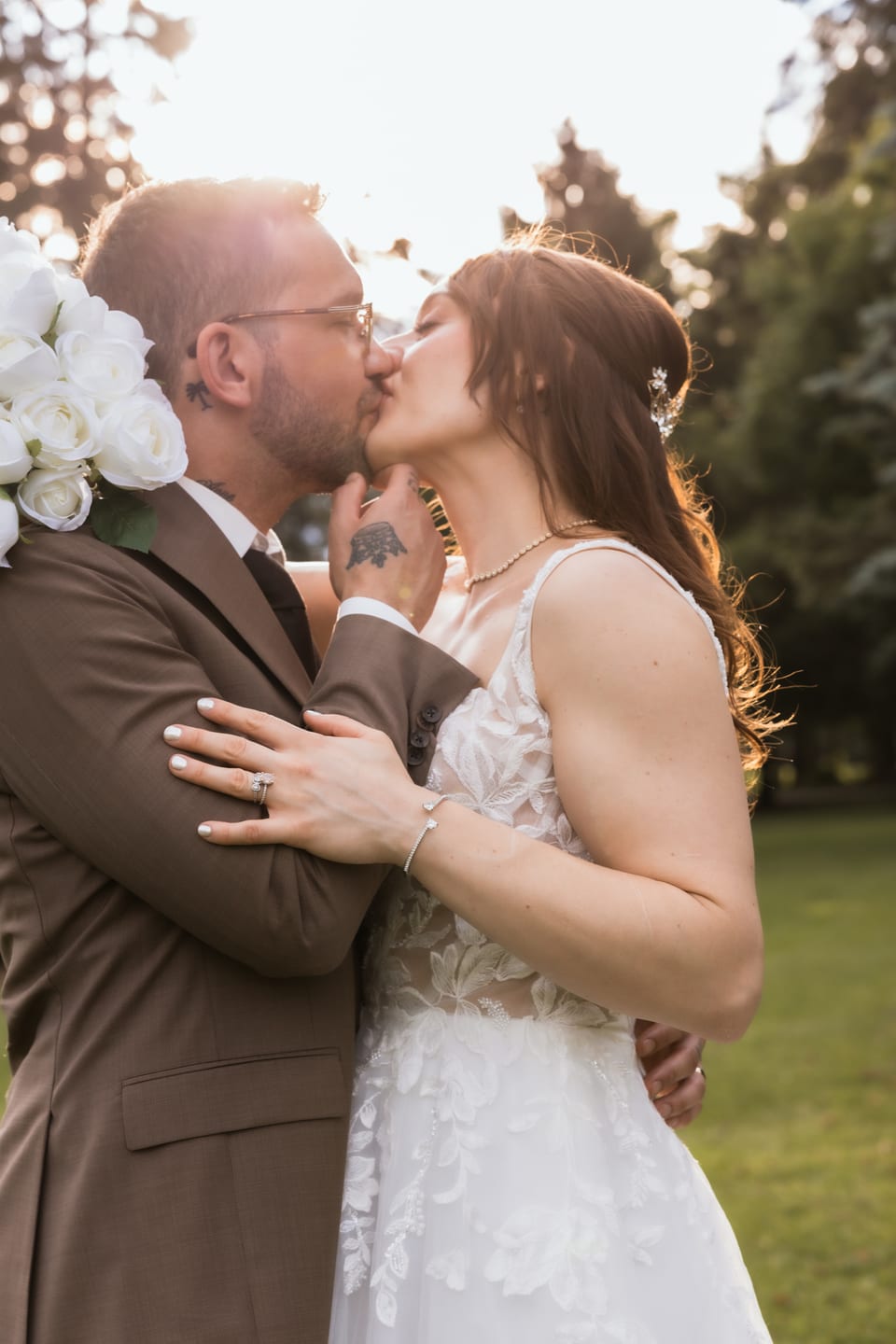 Bride and groom kiss in warm sunset light, her hand resting on his arm, bouquet just in frame.