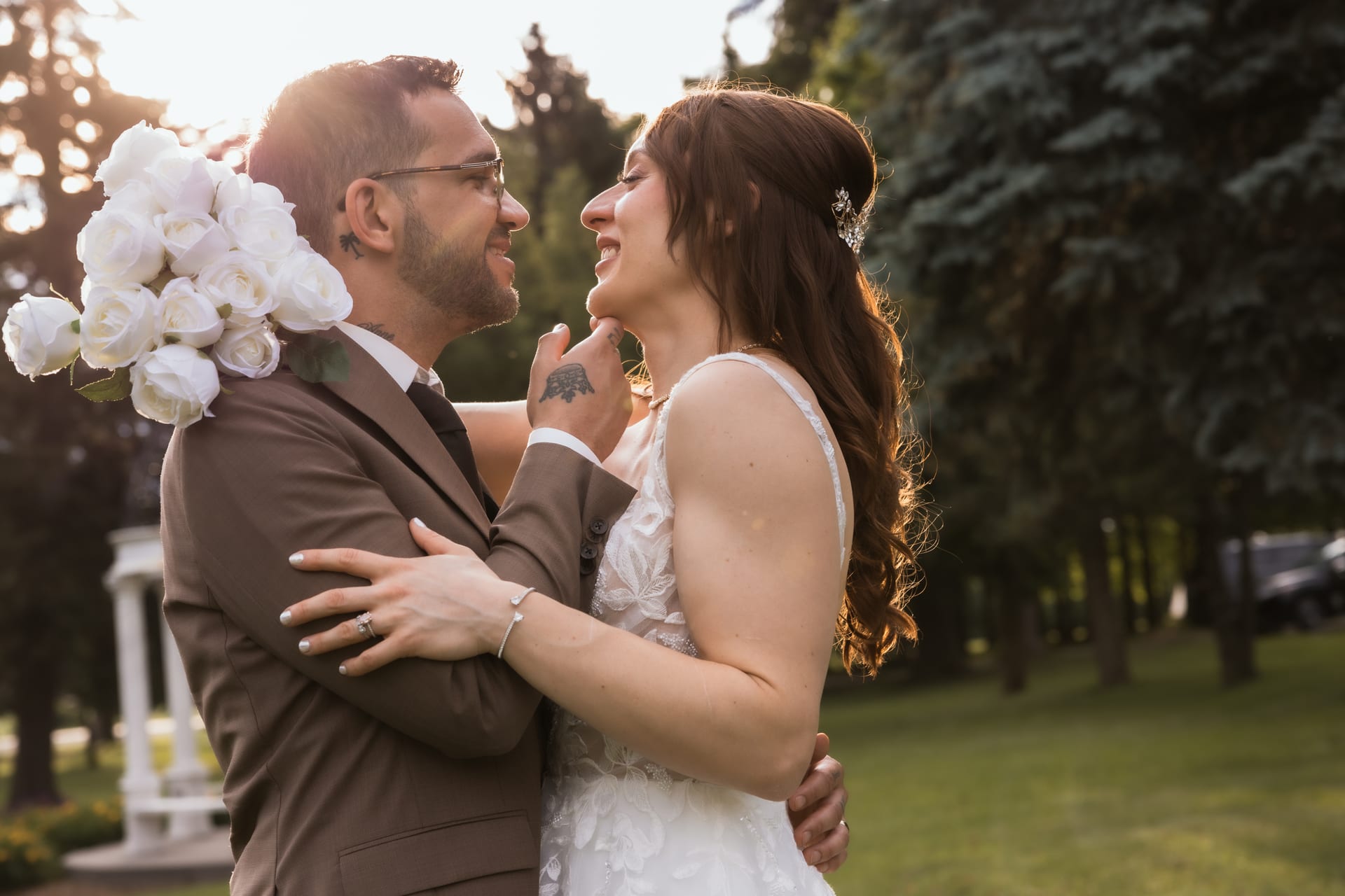 Bride and groom smile nose-to-nose in golden-hour light on the lawn, her bouquet resting on his shoulder.